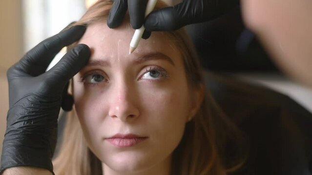 Man's hands in black gloves hold pencil and show on woman's face places for cosmetic injections. Make botox and hyaluronic acid in  beauty salon against wrinkles on forehead.