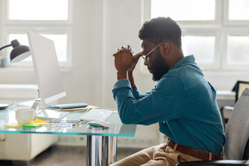 Thoughtful black male entrepreneur in glasses sitting at desk in front of computer, working online...