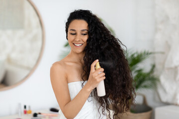 Happy glad millennial caucasian brunette lady in towel applies spray on long curly hair, enjoy treatment - obrazy, fototapety, plakaty