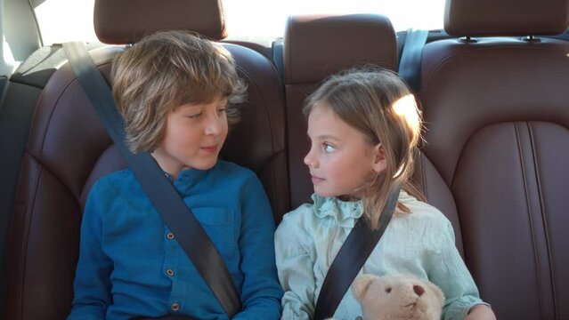 Two little cute children sitting in back seat of car while traveling using tablet smiling and talking. Beautiful blonde boy and lovely pretty girl fastened with safety belts. Car trip concept.