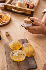 partial view of african american woman holding cosmetic cream near handmade soap on wooden chopping board.