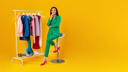 Style assistant. Young lady sitting near clothing rack with colorful clothes, yellow background,...