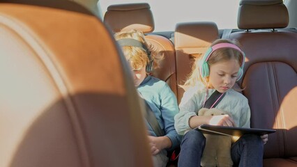 Little cute girl sitting on back seat of car during family car trip with blonde lovely older brother playing games on tablets wearing headphones and listening to music. Safety belts concept.