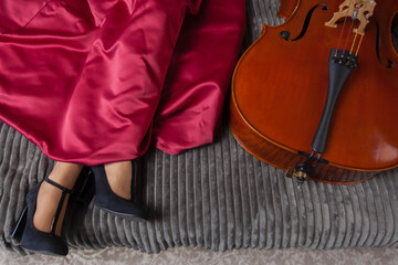 Obraz premium On the bed, woman's legs in a red dress, black high-heeled shoes, and a cello next to her.