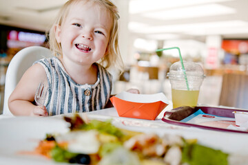 Little girl eats in a cafe. A cheerful child makes faces in a cafe. Some fast food with the family. The cute girl does not want to eat and indulges