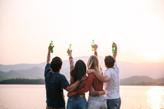 Group of people clinking bottle of beer into the sky while camping outdoors with beautiful sunset view, Happy people on vacation summer.