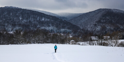 Hiker girl walks along snowy field with mighty mountains in background; hiking in cold winter weather
