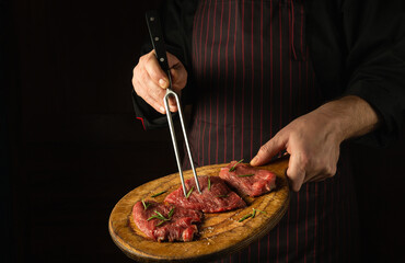 The cook holds a fork in his hand and a cutting board with beef steaks before barbecue. Black space for restaurant recipe or hotel menu.