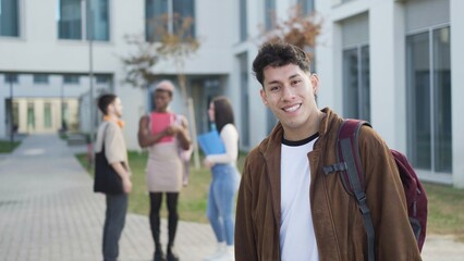 Positive young male student in casual clothes with backpack smiling and looking at camera while standing against classmates near university building