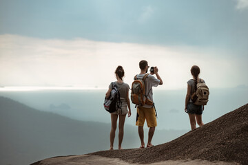 Three young tourists with backpacks stands on mountain top and looks at sea view - obrazy, fototapety, plakaty