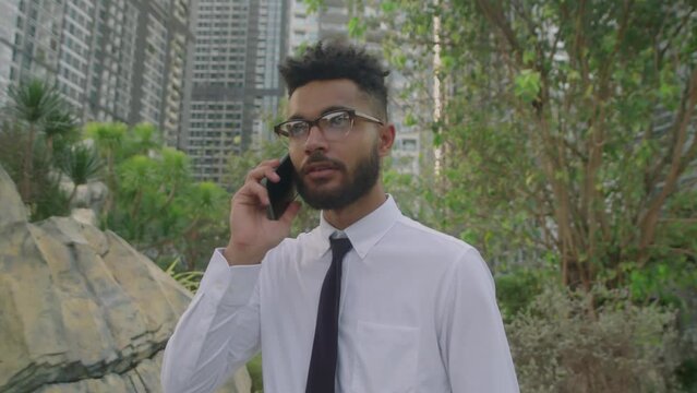 Medium Shot Of Young Businessman In Formalwear Answering Phone Call And Talking While Walking Outside In Park In City