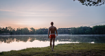 Good looking man in swim shorts going to the lake through the grass and looking at the water