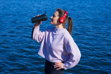 A young athletic woman drinks water after a workout outside.The athlete listens to music with headphones