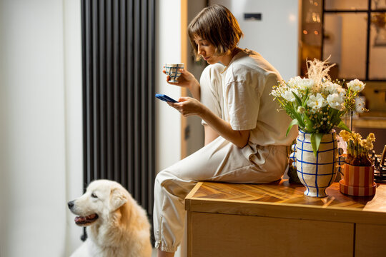 Young woman uses smartphone and drinks a coffee while sitting with her dog on kitchen at home. Concept of morning routine and domestic lifestyle