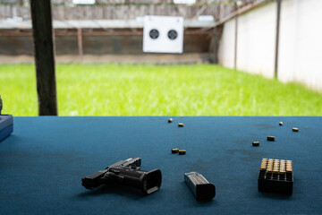 Selective focus of a black pistol with an empty removed magazine, and ammunition in a bullet box, lying together near empty brass shells on the carpet floor with a blurred shooting range in background