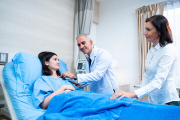 A smiling mid-adult Caucasian male doctor standing using a stethoscope to examine the symptoms of young Caucasian female patient laying on bed with her mother cheering by the bedside in the hospital.