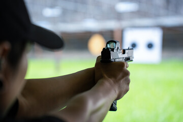 Selective focus of hands of an Asian man wearing a cap, holding a black pistol with straight arms aiming at the front with a blurred paper targets on green grass in the background at shooting range.