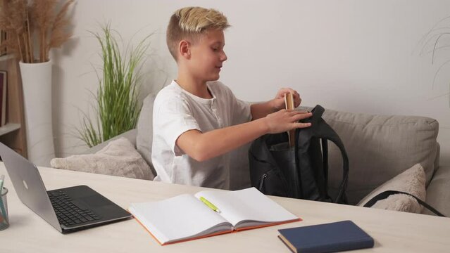 Homework study. Smiling boy. Preparing school. Happy casual teenager kid getting books and work stuff from backpack sitting sofa light room interior.