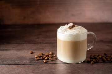 Glass cup of layered coffee drink with milk foam, coffee beans over wooden background, copy space