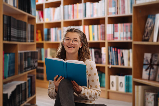 Teen girl among a pile of books. A young girl wearing glasses reads a book with shelves in the background. She is surrounded by stacks of books. Book day.