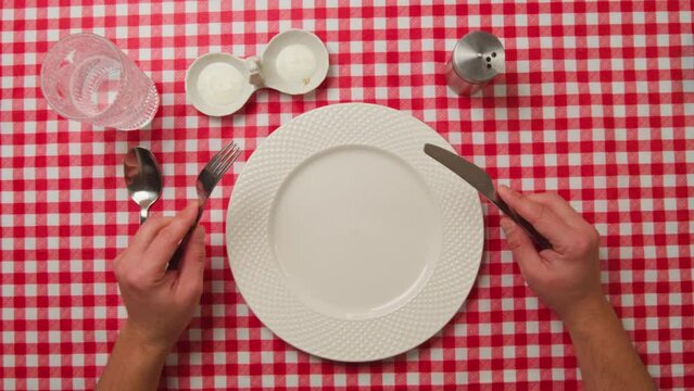 Top view of male hands with cutlery and empty plate on red tablecloth background. Close-up nervous hungry male hands at dinner table holding fork and knife over empty flat white plate