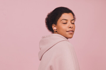 Obraz na płótnie Canvas Stylish fashion african model in wireless earbuds standing with closed eyes on pink pastel background. Lifestyle concept in minimal style and copy space.