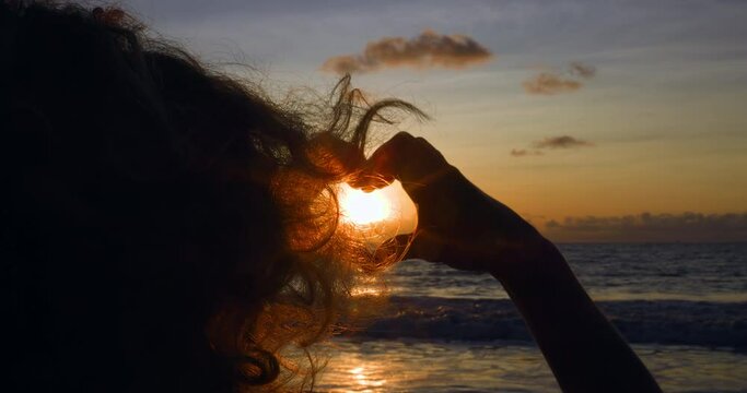 Woman shows heart with hands and watching setting sun on sea. Last day of vacation, memories of which will remain in memory for long time. Woman says goodbye to sea at sunset, showing heart with hands