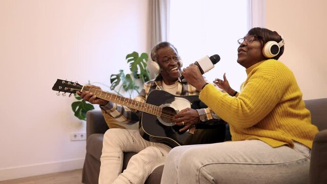 Happy active senior couple play and dance and sing on the couch at home in headphones listening to music
