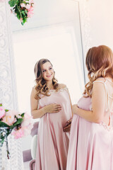 Fototapeta premium a happy pregnant woman in a delicate pink dress looks in the mirror, holding her belly. Surrogate motherhood
