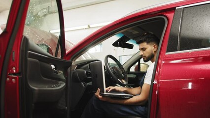 Bearded male mechanic sitting inside car using laptop, recording automobile engine checks collect detailed information during his work on car workshop. Service maintenance during engine repair.