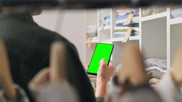 Clothes on a hanger, Active woman Attentively holds the phone looks into the green screen of the smartphone. Stack of seasonal clothes, fashionista girl chooses a dress in a clothing warehouse

