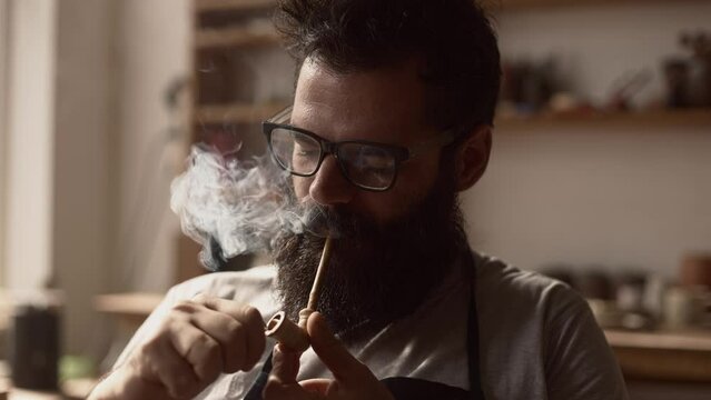 Bearded man in glasses potter professional lights a pipe smokes and drinks coffee during a break from work in his own workshop