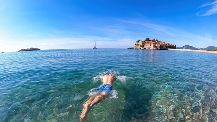 Man bathing in crystal clear water at sand beach next to idyllic island Sveti Stefan, Budva Riviera, Adriatic Mediterranean Sea, Montenegro, Europe. Summer vacation in luxury hotel resort at seaside