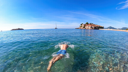 Man bathing in crystal clear water at sand beach next to idyllic island Sveti Stefan, Budva...