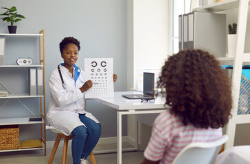 Child is at ophthalmologist's appointment. Female optometrist examines vision of child girl patient using vision chart in hospital office. African American woman doing medical examination of child.