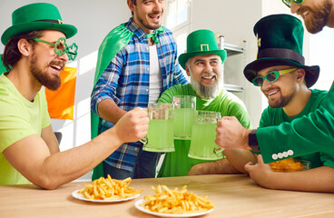 Happy friends having fun at Patrick's Day party. Group of joyful Irish men in green hats and clover glasses sitting at table with French fries, saying toast, clinking big mugs, and drinking green beer