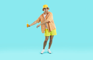 Happy young Caucasian man tourist performs dance with glass of juice in hands enjoying trip to tropical island or vacation at resort dressed in summer casual clothes, stands in turquoise studio