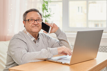 Cheerful senior man has phone conversation, chatting on the smartphone working with a laptop remotely from home.