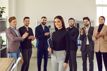 Portrait of successful female company leader or business team manager at work. Happy beautiful young woman with folder in hand standing in modern office, with group of employees clapping in background