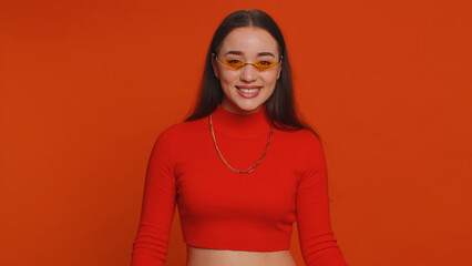 Fototapeta premium Portrait of happy pretty young woman in crop top wearing sunglasses, looking at camera with toothy charming smile, flirting, expressing optimism. Millennial girl isolated on red studio background