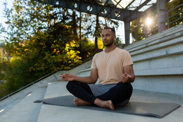 A young African American man is sitting on a mat in a stadium in the lotus position and doing yoga....