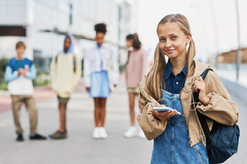 Blond pretty schoolgirl with backpack and smartphone looking at camera while standing against group of intercultural friends outdoor - obrazy, fototapety, plakaty
