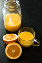 A dark and moody scene of bright orange fruit juice on a black cement surface featuring fresh sliced oranges. The juice is in a large mason jar and also in a glass clear mug with a handle.