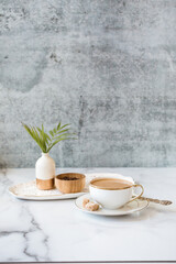 Small bud vase with a palm leaf sits next to a ramekin of roasted coffee beans and sits on a white tray with gold. It also features coffee in a teacup with cream and sugar cubes.