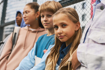 Cute blond schoolgirl in casualwear looking at camera among her friends or classmates while...