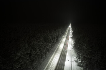An atmospheric drone photo of a winter road winding through a snow-covered forest at night. The trees are adorned with a delicate layer of hoarfrost, creating a magical winter wonderland