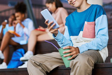 Close-up of adolescent boy in casualwear texting in mobile phone and having soda while sitting in front of camera against his friends