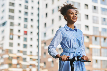 Happy adolescent African American schoolgirl in blue casual dress looking forwards while riding...