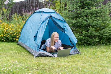 A young girl lying on the grass in the tourist tent with a laptop and communicates through a video call.