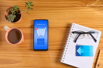Top view of mobile phone on wooden table with interface of online store on the screen, shopping cart icon and buttons to view cart and checkout. With credit card and accessories on the table.